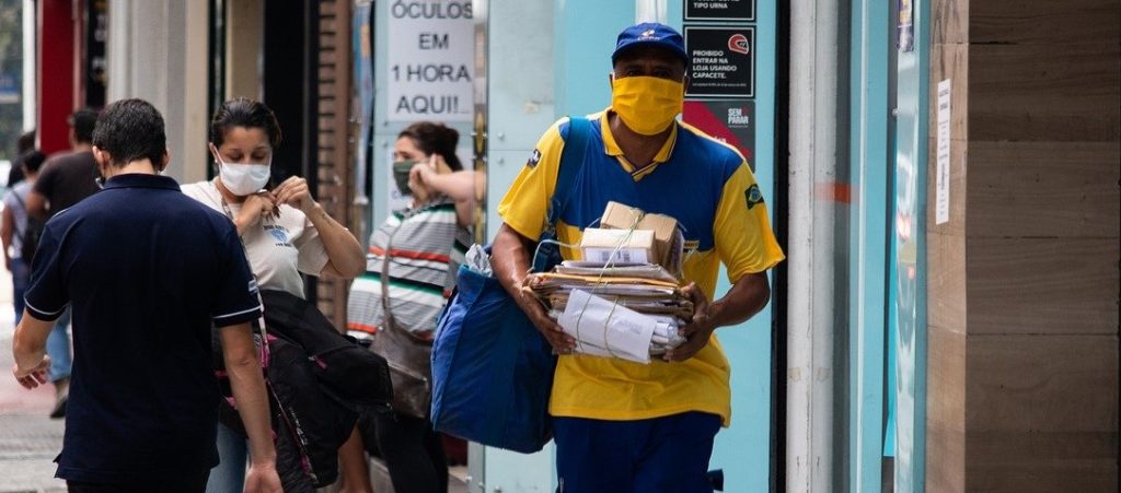 People are in the streets during COVID-19 pandemic wearing masks; a postman is in the front of the picture with small packages in his hands.